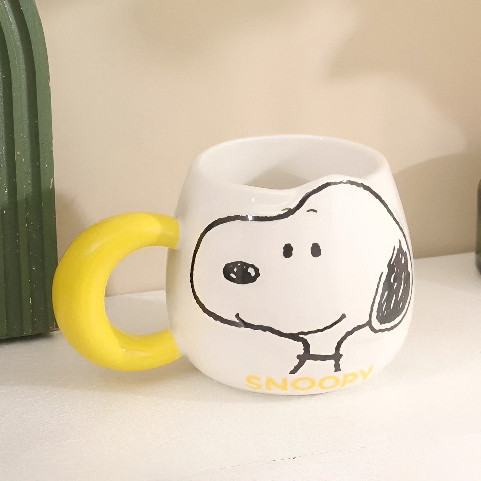 Bijela keramička šolja s žutim držačem i crnim crtežom snoopyja s natpisom snoopy u žutom na dnu