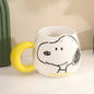 Bijela keramička šolja s žutim držačem i crnim crtežom snoopyja s natpisom snoopy u žutom na dnu